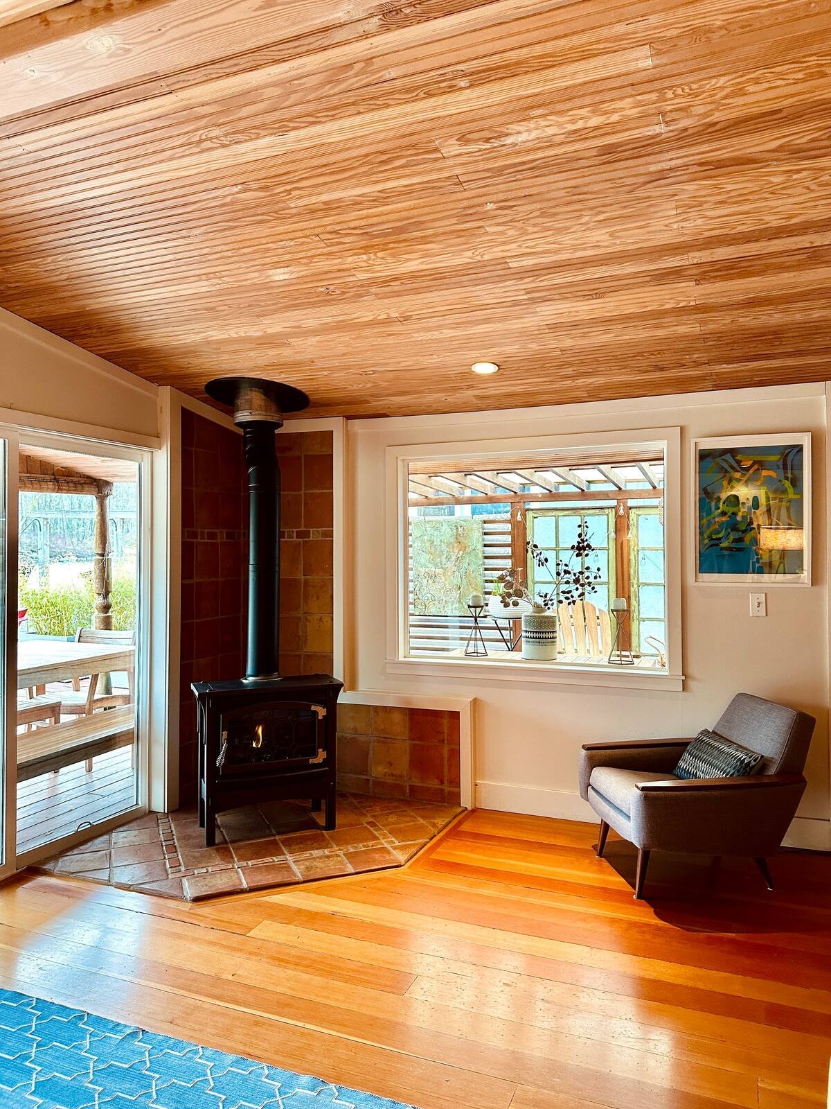 Wood stove corner with fire, leather chair, cedar ceiling, and glass doors to porch